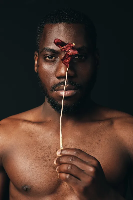 Black man with bare chest holding dried red flower in front of face, creative conceptual portrait with dark background and artistic visual style, African ethnicity.