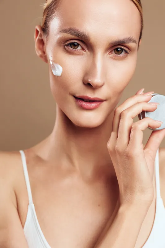 Young woman applying facial cream on cheek, skincare routine, natural makeup, clear skin, soft lighting, beauty care concept, neutral background, close up portrait
