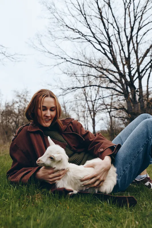 Lifestyle scene with a woman relaxing on grass, holding a goat kid, smiling, outdoors in a park, nature and affection visible, casual cozy attire, calm moment in autumn sunlight