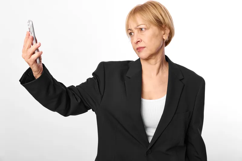 Senior woman in business attire holding smartphone for selfie, surprised expression, studio portrait on white background, communication technology concept, modern work routine