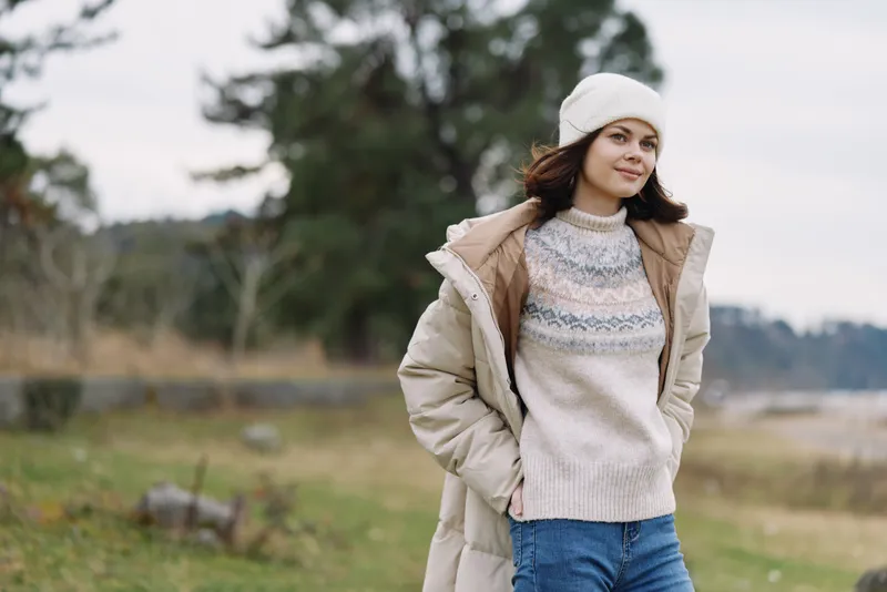 Woman in winter attire outdoors wearing a beige coat knitted sweater and white beanie standing in a rural field with trees behind