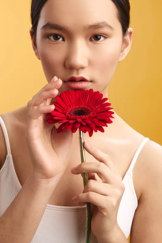 Asian woman holds a vibrant red flower near her face in a studio portrait with a warm yellow backdrop, a calm expression, and soft lighting, conveying beauty, focus, and contemporary elegance.