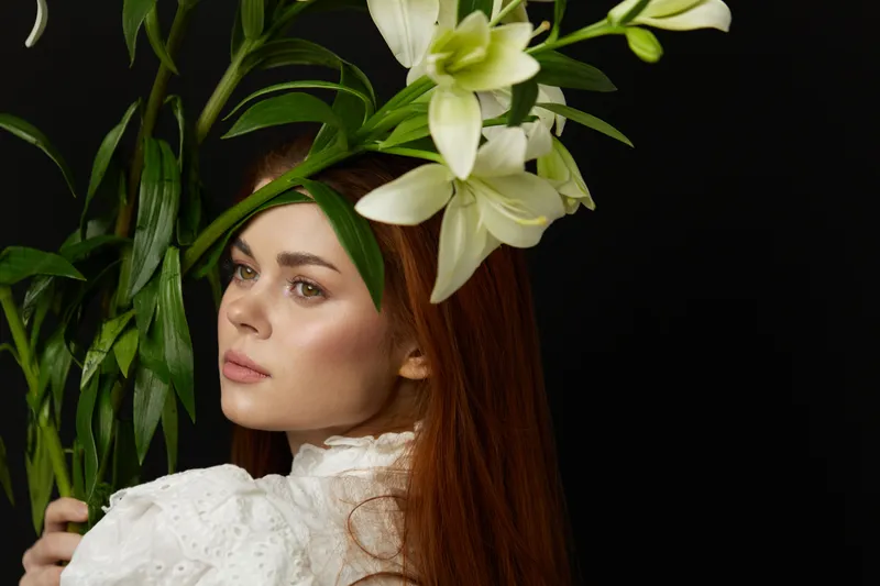 Young woman with long red hair, wearing a white vintage dress, holding fresh flowers, featuring a serene expression against a dark background