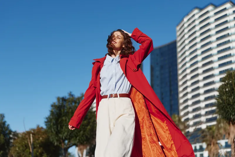 Young woman in red coat and white pants walks confidently outdoors in urban environment with modern buildings and trees under clear blue sky.