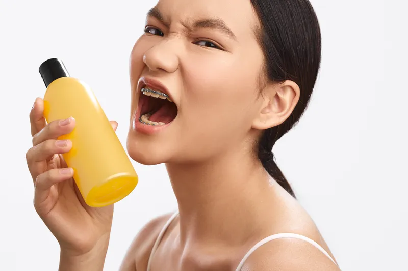 Asian woman holds a yellow bottle close to her face, smiling with enthusiasm in a bright studio. The image conveys freshness, self care, and a vibrant home beauty routine.