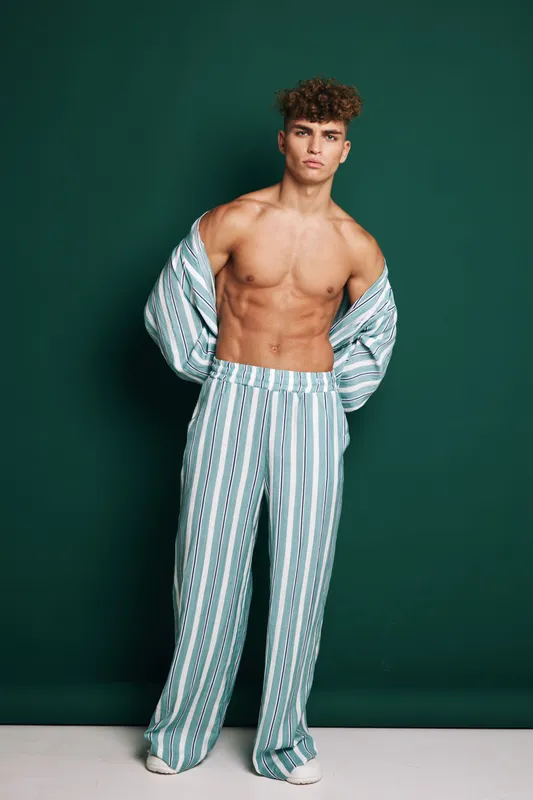 Young man with muscular body posing shirtless wearing striped pants and open shirt against green background. Fashion model with curly hair showing confident expression in studio.