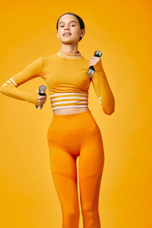 Female fitness athlete in bright orange athleisure holding dumbbells, mid workout pose against a vivid backdrop, showcasing strength, balance, and athletic confidence during training.