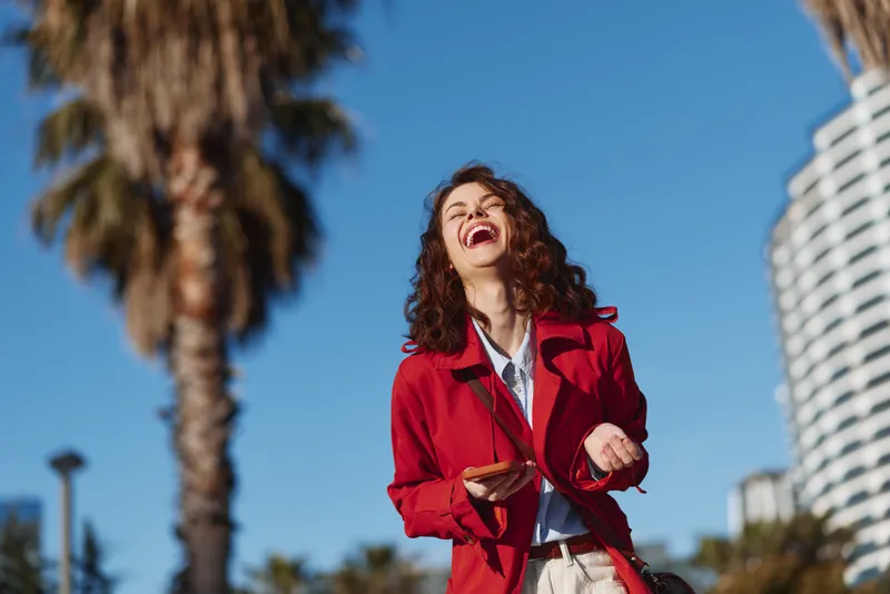 Laughing young woman in a bright red jacket stands outdoors in a sunny cityscape with palm trees behind, capturing a carefree moment.