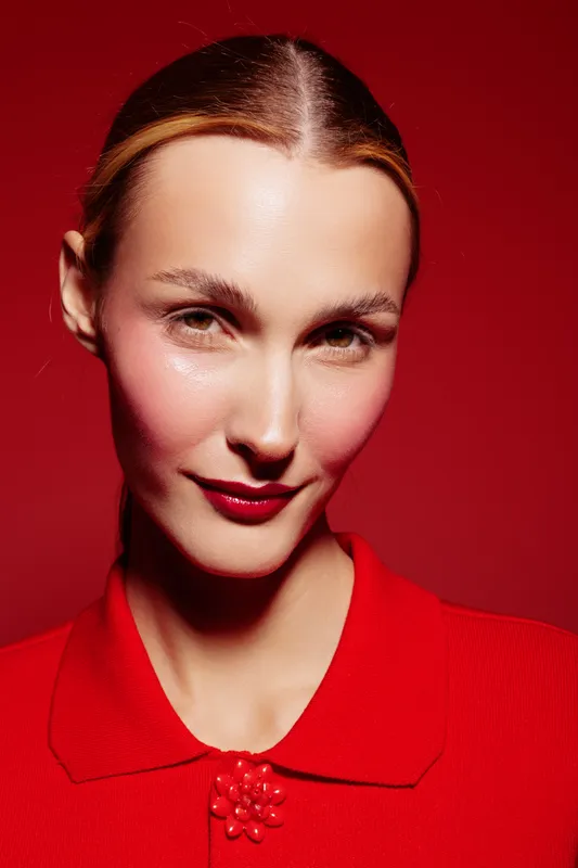Portrait of confident woman with slicked hair wearing red blouse against red background. Studio lighting highlights facial features and makeup. Concept of fashion and elegance.