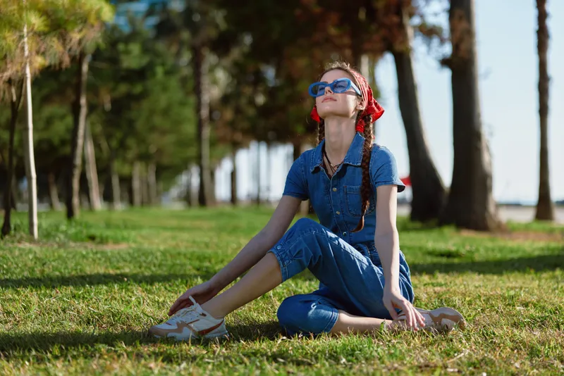 young woman sitting on grass outdoors wearing blue denim jumpsuit and sunglasses enjoying warm sunny day in green park environment