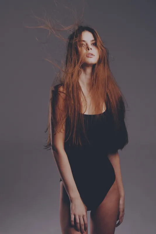 Young woman with long red hair in black bodysuit posing against gray background, fashion concept, elegant style, studio lighting