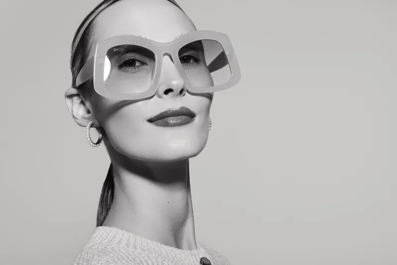 Portrait of a confident Caucasian woman with pale skin wearing oversized square sunglasses and a light sweater. Studio shot in black and white with smooth lighting.