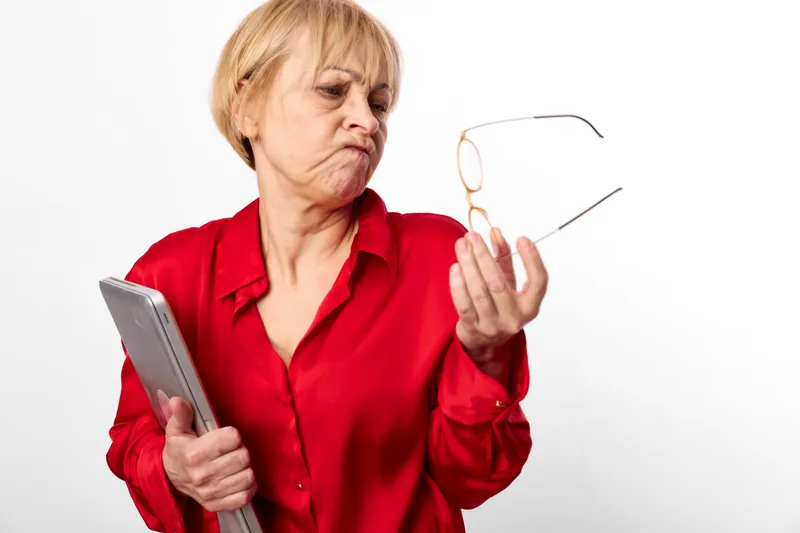 Elderly woman red shirt glasses reading document, annoyed expression, holds eyeglasses in one hand, isolated on white background, studio portrait, copy space, care