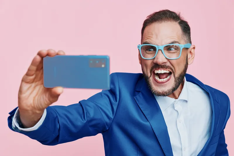 A professional man in a blue suit takes a bold selfie against a solid pink background, showcasing high energy, confidence, and playful excitement.