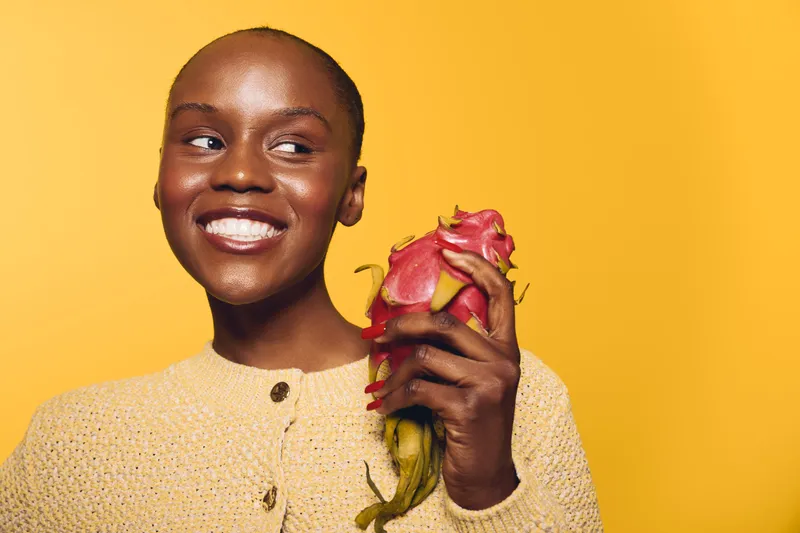 Smiling black woman with dark skin holds a dragon fruit near her face against yellow background. Natural beauty concept with fresh fruit and positive expression in studio lighting.