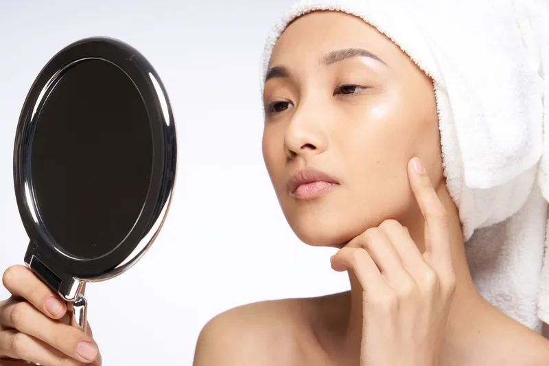 Азиатская женщина с обнаженными плечами касается рукой своего лица и смотрится в зеркало