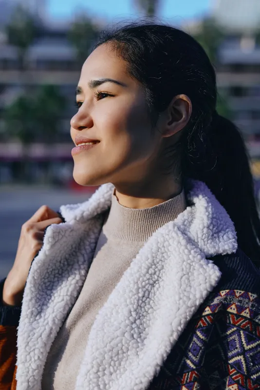 Woman in a shearling coat outdoors, bathed in warm sunlight, gazing to the side with a calm smile, textured fabric, casual fashion, serene mood in a daylit urban setting.