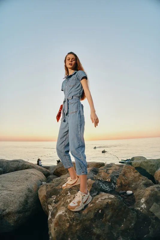 Lifestyle woman in modern boho-western denim street style standing on rocks by the sea during golden hour, film color warm tones, casual urban fashion vibe