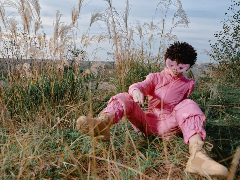 Woman in pink jumpsuit sitting among tall grasses outdoors, relaxed pose with boots, nature around and an expansive landscape under a clear sky, peaceful mood and open space