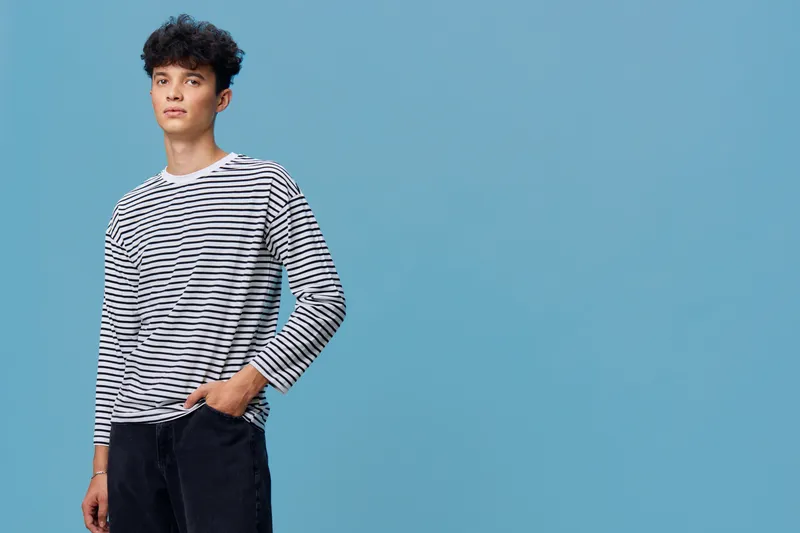 young man standing with hand in pocket wearing striped long-sleeve shirt against blue background in casual style