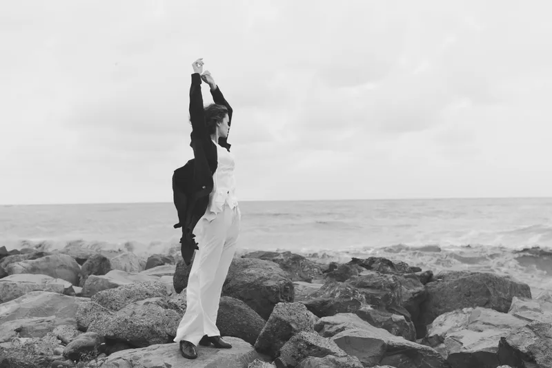 Couple in formal attire dancing on a rocky shoreline by the sea, black and white image capturing romance, celebration, and wind swept scenery with crashing waves behind