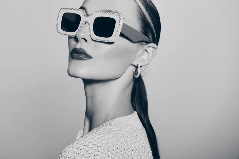 Stylish Caucasian woman with light skin wearing large square sunglasses and textured sweater poses confidently in monochrome studio. Fashion and elegance concept with modern accessories.