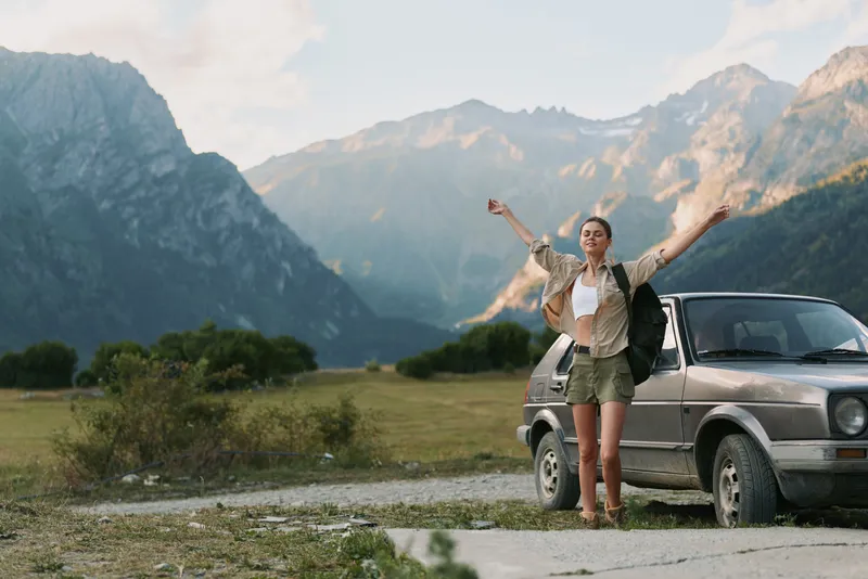 Young woman with long hair and backpack stands next to a car in a mountain valley raising her arms in joy during a hiking trip.