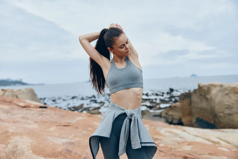A young woman of mixed descent stretches on rocky coastline, wearing a gray sports bra and black leggings, embodying fitness and a healthy lifestyle by the sea