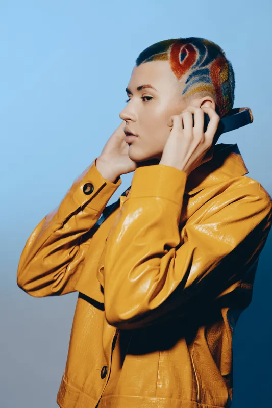 woman with colorful patterned shaved hairstyle wearing a shiny yellow jacket holding a phone against a blue background, stylish and modern look