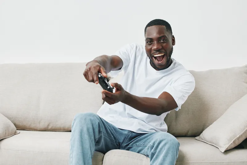 Happy young Black man playing video games on the couch with joy, wearing casual white t shirt and jeans, indoor setting in light colors