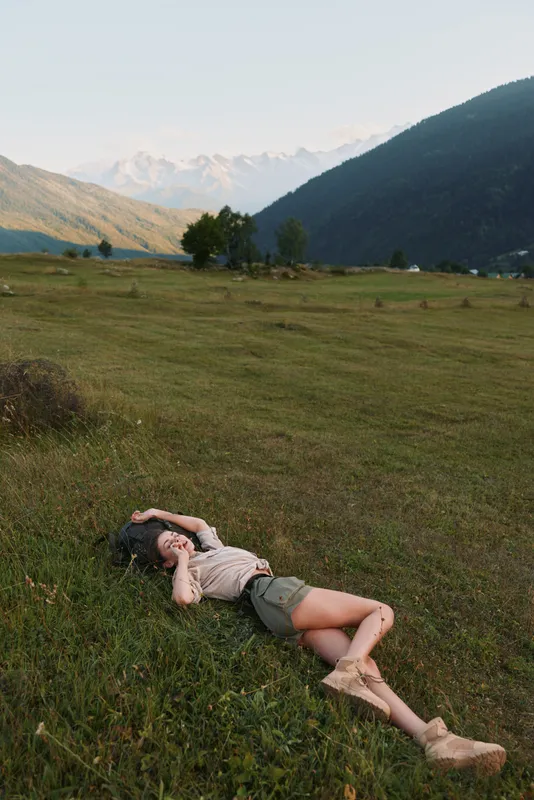 Cheerful woman lying in a grassy meadow outdoors, sunlit mountains in the background, relaxed pose, gentle breeze, natural colors, carefree lifestyle in bright daylight