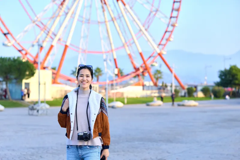 Female traveler with camera stands in an open amusement park, smiling, with a dramatic ferris wheel backdrop, casual jacket, denim, sunny day, leisure photography setup, capturing joyful moments