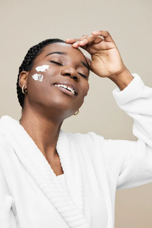 Smiling black woman applying facial cream, showcasing a skincare routine, in cozy white robe against a neutral background highlighting beauty and self care