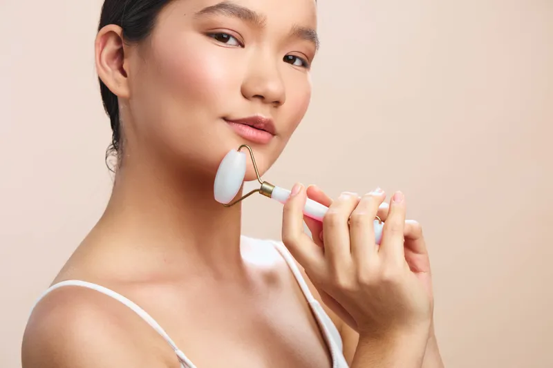 asian woman applying skincare tool in a bright modern room, she smiles softly, relaxed and confident, showcasing a gentle beauty routine and personal care moment.