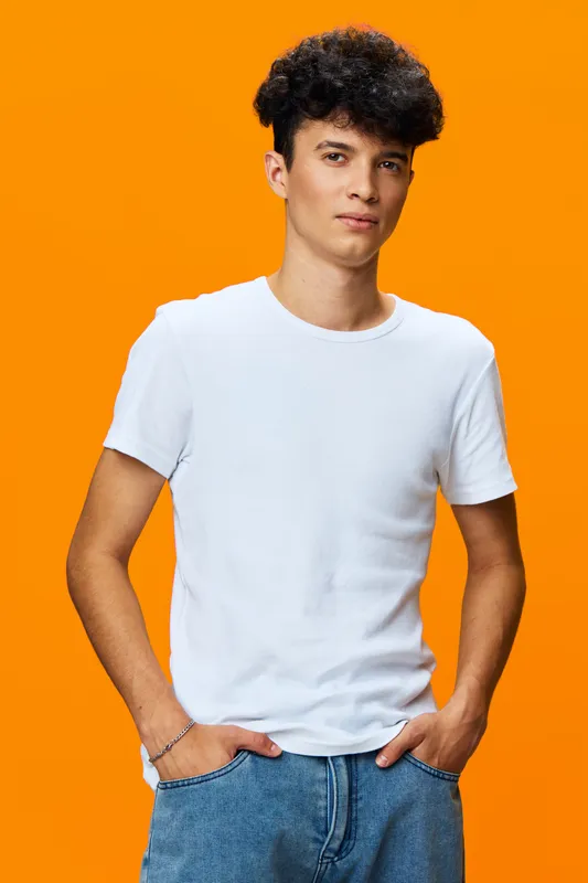 Young man in white t-shirt and blue jeans standing confidently against bright orange background with casual posture and relaxed expression.
