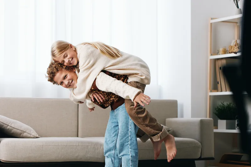 Playful couple shares a cozy piggyback moment in a bright living room, capturing joyful affection, casual attire, soft lighting, and a relaxed home ambiance.