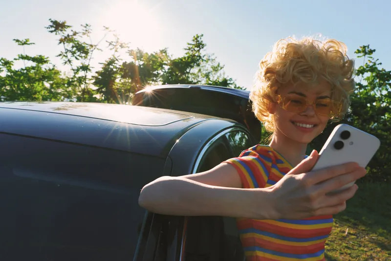 A cheerful child takes a selfie beside a car in bright sunlight, striped shirt and curly hair, capturing playful outdoor moments. Concept: carefree youth, summer lifestyle, candid portrait.