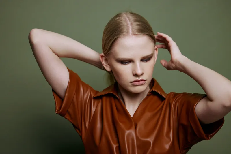 Модная стильная женщина, касаясь волос, позирует изолированно в коричневой палитре: кожаное платье и тренчкот на фоне оливково-зеленой стены
