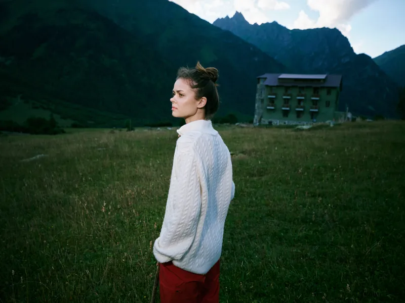 A woman in a white knit sweater stands in a grassy field with distant mountains and a single rural building, creating a serene rural landscape and calm mood.