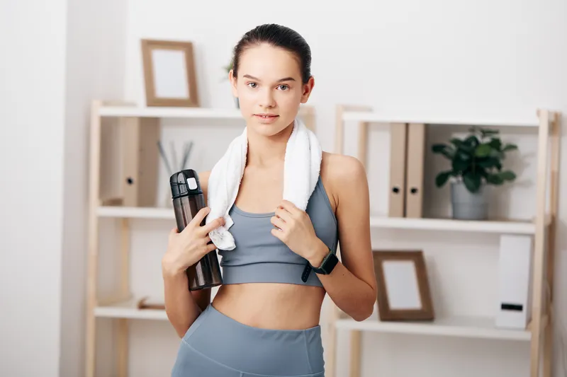 Female fitness enthusiast in gym attire with a towel and water bottle, confident pose during workout amid a clean backdrop of shelves and neutral tones.