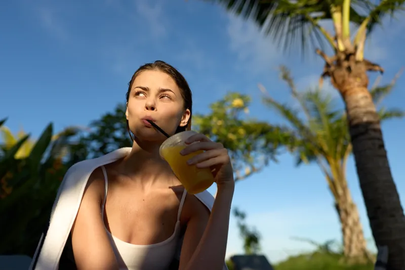 Smiling woman outdoors sipping a yellow beverage, casual summer scene with palm trees, bright sunlight. Focus on refreshment, wellness, and self care in a relaxed lifestyle moment.
