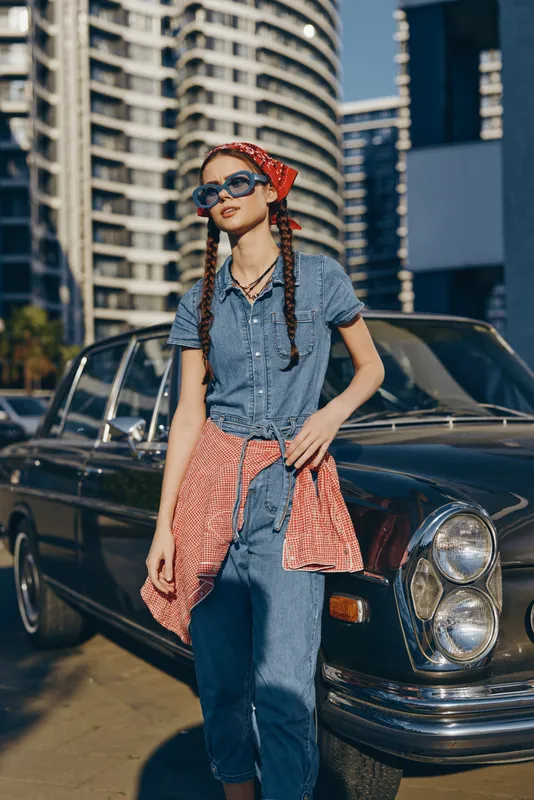 young woman with braids and sunglasses standing near vintage black car wearing denim outfit and red checkered shirt tied around waist in urban setting