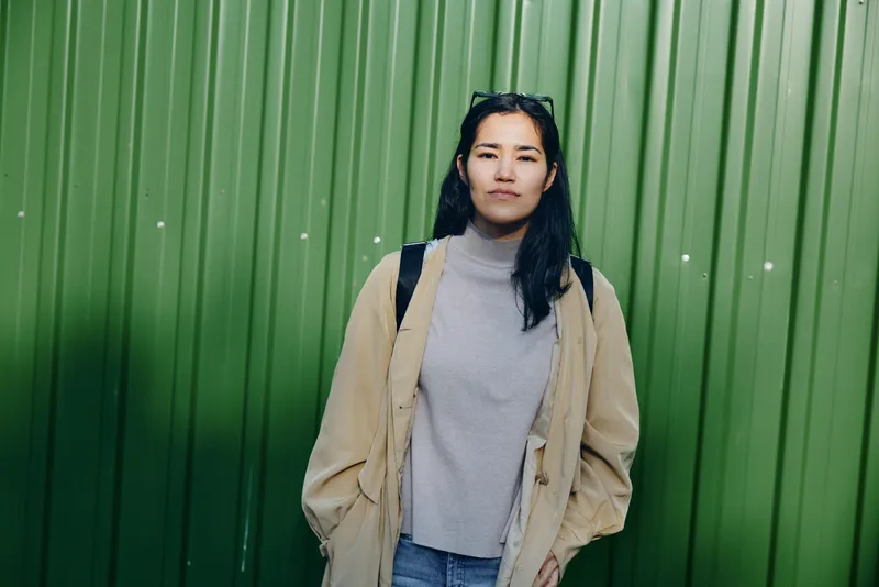 Woman in beige cardigan and gray turtleneck stands on a street against a green wall, casual fashion pose with backpack, urban style and relaxed mood captured outdoors.
