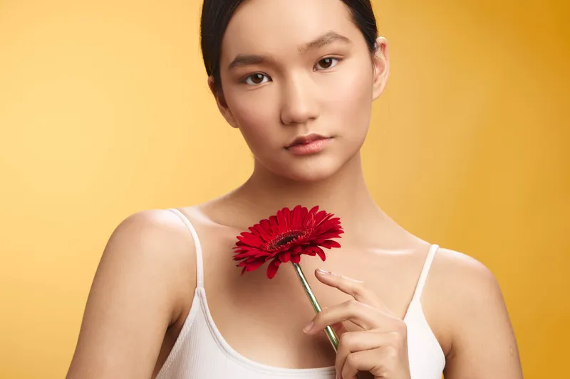 Asian woman gently holds a bright red flower against a warm yellow backdrop, presenting a calm and graceful portrait with natural makeup and a focused, serene expression.