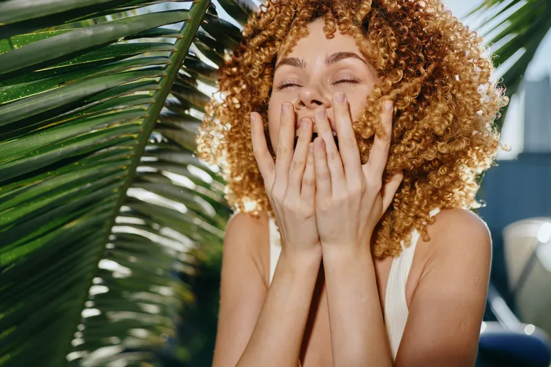 smiling woman with curly hair enjoying sunshine outdoors surrounded by tropical green leaves, natural light, and fresh summer vibes.