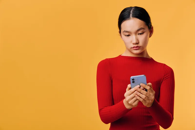 Young woman in red sweater standing against orange background looking down at smartphone in hands for texting communication and social media using technology device.
