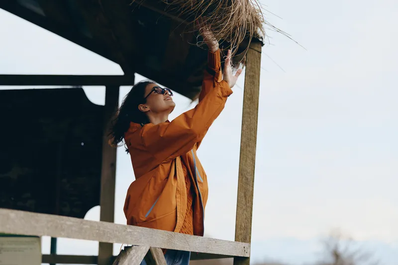 Woman lifestyle outdoors in orange jacket, standing by wooden railing, looking upward in bright sunlight, blue sky background, casual leisure mood, natural light, calm expression