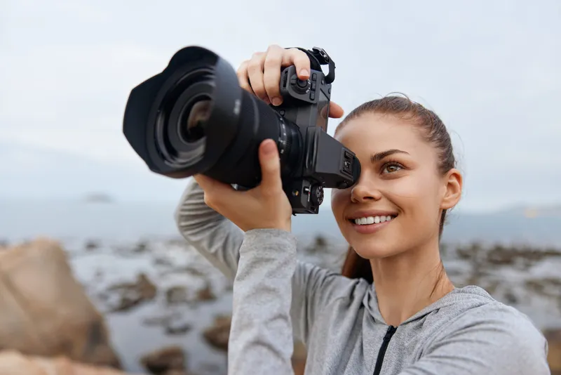 Young woman smiling while taking photos with a camera by the seaside, showcasing joy and creativity Perfect for lifestyle and photography themes