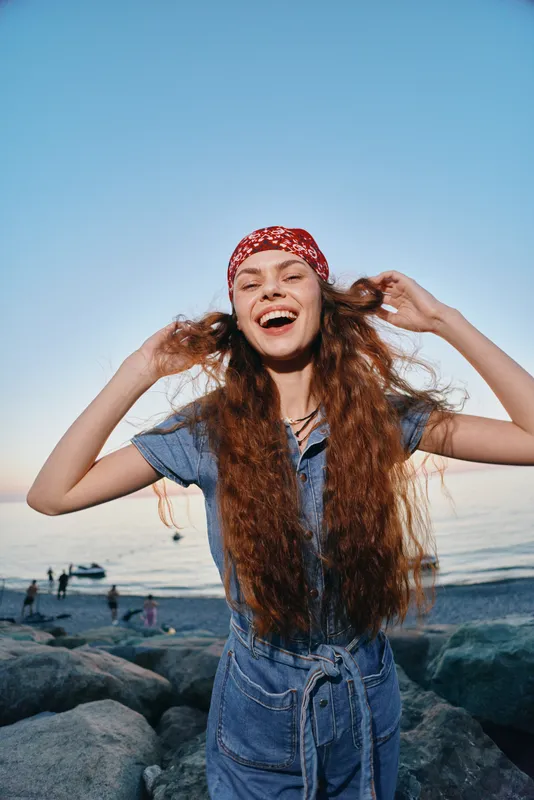 Lifestyle woman in modern boho-western denim street style laughing joyfully with long hair and red bandana by the sea in warm film color tones