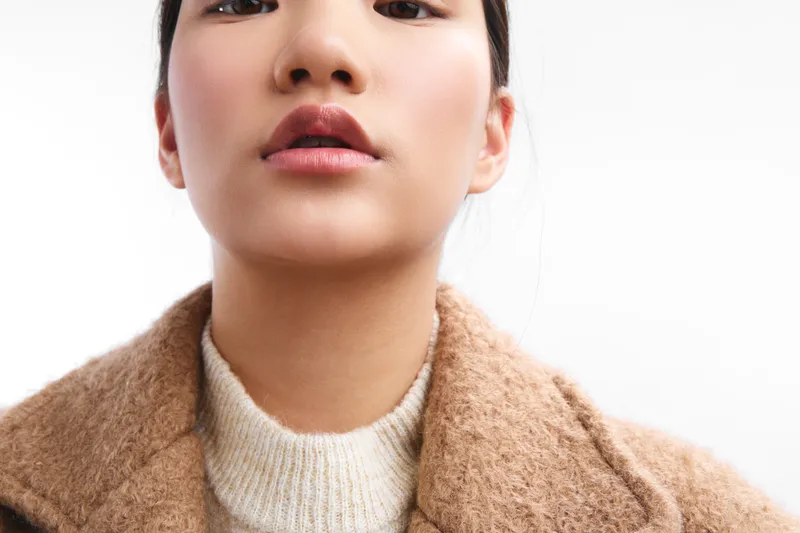 Asian woman in a warm coat stares softly at the camera, exuding calm confidence and thoughtful mood. A close up emphasizes texture, tone, and natural elegance, capturing subtle details of expression